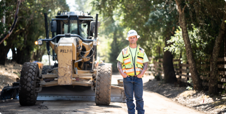 Dirt World worker standing in front of a motor grader